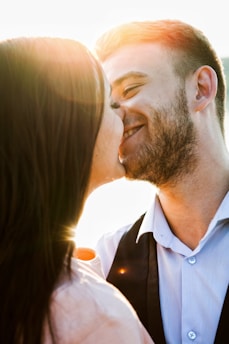 A couple shares a romantic moment as they lean in for a kiss, illuminated by warm sunlight in the background, casting a soft glow around them.