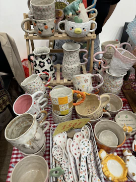 Various handcrafted ceramic cups, mugs, and spoons are displayed on a checkered tablecloth. The ceramics feature colorful designs, including floral patterns, polka dots, and whimsical shapes. Some cups have handles in unique shapes, and a small price tag is visible among the items.