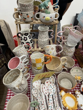 Various handcrafted ceramic cups, mugs, and spoons are displayed on a checkered tablecloth. The ceramics feature colorful designs, including floral patterns, polka dots, and whimsical shapes. Some cups have handles in unique shapes, and a small price tag is visible among the items.