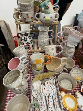Various handcrafted ceramic cups, mugs, and spoons are displayed on a checkered tablecloth. The ceramics feature colorful designs, including floral patterns, polka dots, and whimsical shapes. Some cups have handles in unique shapes, and a small price tag is visible among the items.