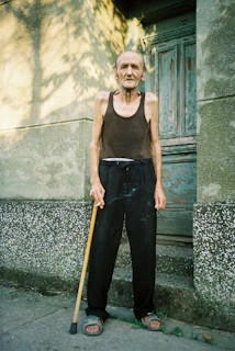 An elderly man, wearing a black sleeveless shirt and black pants, is standing on a concrete surface near a textured wall and an old wooden door. He is holding a walking cane in one hand and appears to be outdoors in a shaded area with sunlight filtering through.