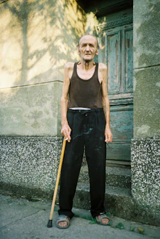 An elderly man, wearing a black sleeveless shirt and black pants, is standing on a concrete surface near a textured wall and an old wooden door. He is holding a walking cane in one hand and appears to be outdoors in a shaded area with sunlight filtering through.