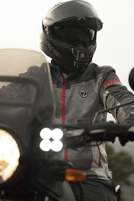 A motorcyclist wearing a full-face helmet with a mounted camera on it. The person is dressed in a protective grey and red jacket. The motorcycle's headlights are on, illuminating the foreground. The background is blurred, indicating motion or focus on the rider.