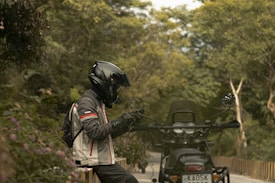 A motorcyclist in protective gear is seated on a motorcycle parked along a tree-lined road. The rider is wearing a black helmet and a jacket with red accents. The surrounding area is lush with greenery, including trees and flowering plants. The atmosphere suggests a serene and tranquil outdoor setting.