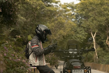A motorcyclist in protective gear is seated on a motorcycle parked along a tree-lined road. The rider is wearing a black helmet and a jacket with red accents. The surrounding area is lush with greenery, including trees and flowering plants. The atmosphere suggests a serene and tranquil outdoor setting.