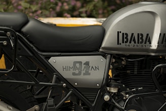Close-up of the side of a motorcycle with a focus on the seat and fuel tank. The tank is metallic grey with text and coordinates, and the side panel is labeled 'HIMALAYAN 91.' The seat is upholstered in black leather, and various metal components of the motorcycle's frame and engine are visible.