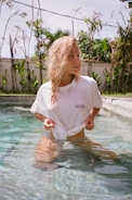 A person with curly blonde hair is standing in a shallow pool of water. They are wearing a white T-shirt with the words 'Emotionally Unavailable' printed on it. The background shows a garden with greenery, including palm trees and other plants, under a bright sky.