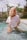 A person with curly blonde hair is standing in a shallow pool of water. They are wearing a white T-shirt with the words 'Emotionally Unavailable' printed on it. The background shows a garden with greenery, including palm trees and other plants, under a bright sky.