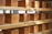 Several loaves of bread are neatly arranged on wooden shelves. The loaves are uniformly shaped and have a golden-brown crust. Some natural light seems to be entering from a window in the background, illuminating the loaves.