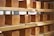 Several loaves of bread are neatly arranged on wooden shelves. The loaves are uniformly shaped and have a golden-brown crust. Some natural light seems to be entering from a window in the background, illuminating the loaves.