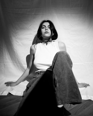 A black and white photograph of a person sitting on the floor against a plain backdrop. The person is wearing a sleeveless top and loose-fitting pants, with one knee raised. The expression conveys a sense of confidence and introspection.