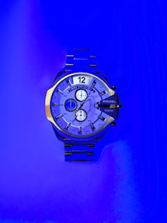 A large wristwatch with a metallic strap is displayed against a vibrant blue background. The watch face is detailed with multiple dials and has a bold design. The watch brand 'Diesel' is visible on the right side of the watch face.
