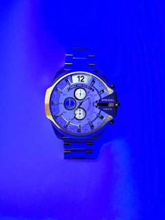 A large wristwatch with a metallic strap is displayed against a vibrant blue background. The watch face is detailed with multiple dials and has a bold design. The watch brand 'Diesel' is visible on the right side of the watch face.