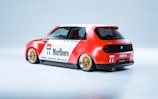 A compact car with a sporty design featuring a red and white color scheme. The car is adorned with racing decals, including the number 77, and logos from brands such as Marlboro, Honda, and Michelin. It has distinctive golden wheels and a streamlined body shape, conveying a sense of speed and style.
