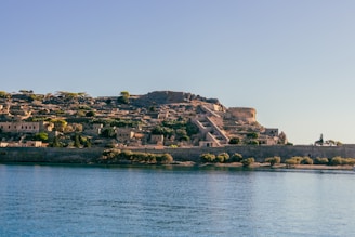 A sunlit, ancient fortress overlooking a calm body of water. The fortress is made of stone with visible structures, pathways, and walls. Vegetation including trees and bushes surrounds the fortress, adding greenery to the scene. The sky is clear and blue, creating a serene atmosphere.