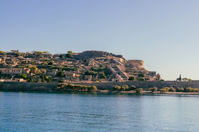 A sunlit, ancient fortress overlooking a calm body of water. The fortress is made of stone with visible structures, pathways, and walls. Vegetation including trees and bushes surrounds the fortress, adding greenery to the scene. The sky is clear and blue, creating a serene atmosphere.