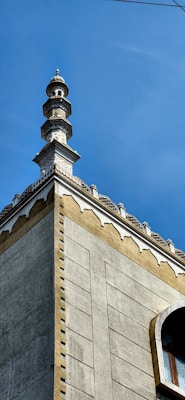 A tall, ornate minaret with intricate architectural details rises against a clear blue sky. The structure features a series of decorative tiers and geometric patterns, topped with a pointed spire. The building's facade is textured in light and dark tones, and a small section of a window is visible.