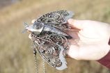An intricately designed piece of jewelry resembling a butterfly is held by a hand. The butterfly features detailed metalwork with swirling patterns and dark spherical stones. The body of the butterfly appears to be made of a smooth, marbled material with a delicate structure. The background is a blurred, grassy field, lending a natural setting to the image.