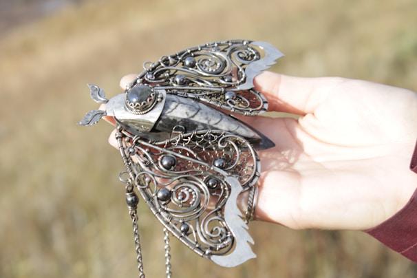 An intricately designed piece of jewelry resembling a butterfly is held by a hand. The butterfly features detailed metalwork with swirling patterns and dark spherical stones. The body of the butterfly appears to be made of a smooth, marbled material with a delicate structure. The background is a blurred, grassy field, lending a natural setting to the image.