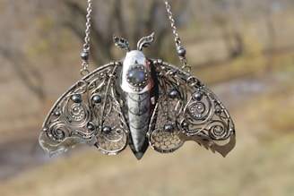 A beautifully crafted pendant designed to resemble a moth, featuring intricate metalwork. The wings are adorned with swirling patterns and spherical embellishments, and a central stone is set into the body of the moth.