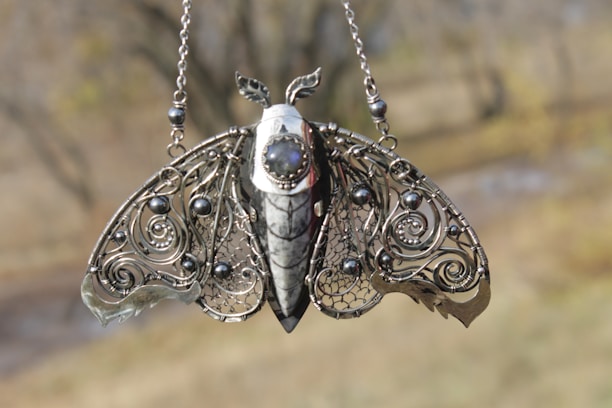 A beautifully crafted pendant designed to resemble a moth, featuring intricate metalwork. The wings are adorned with swirling patterns and spherical embellishments, and a central stone is set into the body of the moth.