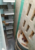 A multi-story building interior with a view of staircases on one side and a curved reception area on the other. The walls are decorated with beige tiles and there are green panels running vertically. A few office chairs and desks are visible in the reception area, and the flooring features dark tiles.