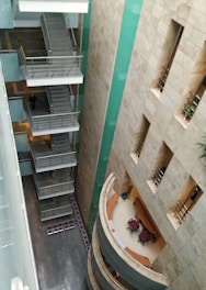A multi-story building interior with a view of staircases on one side and a curved reception area on the other. The walls are decorated with beige tiles and there are green panels running vertically. A few office chairs and desks are visible in the reception area, and the flooring features dark tiles.