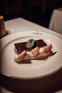 A rectangular chocolate dessert is elegantly presented on a white plate, garnished with fresh mint leaves, dollops of whipped cream, and slices of fresh strawberries.