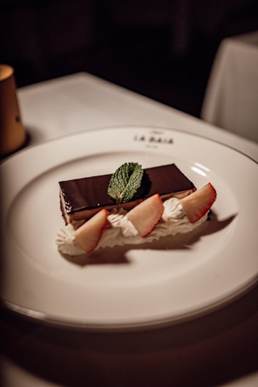 A rectangular chocolate dessert is elegantly presented on a white plate, garnished with fresh mint leaves, dollops of whipped cream, and slices of fresh strawberries.