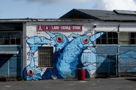 A mural on the side of a brick warehouse building features three large, blue, abstract bird-like creatures with exaggerated eyes. The building facade is old and weathered, with various windows and a central grid-style window incorporated into the mural. Above the mural, there is a sign with a red background and white text in three languages, indicating 'Store' as one of the words.