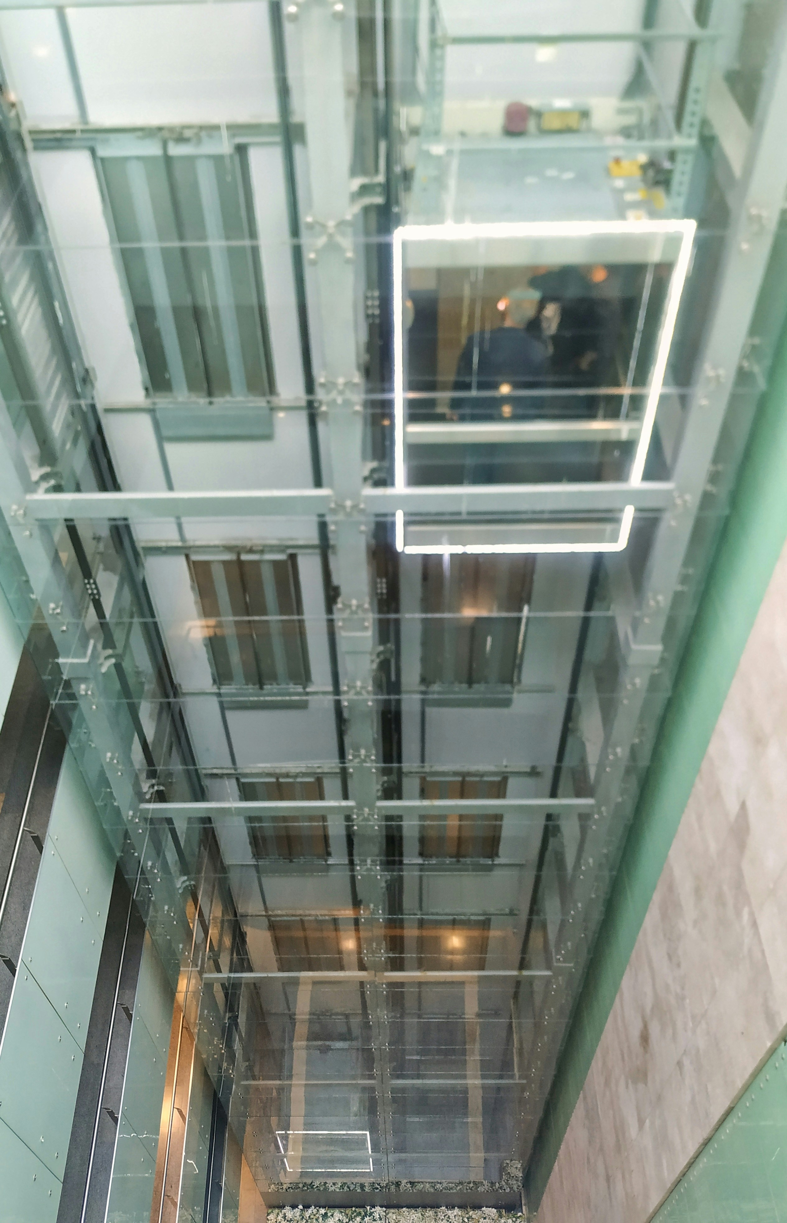 A glass elevator shaft extending vertically with a view upwards. A glass-walled elevator cabin is visible with two people inside. The structure is predominantly metallic and transparent, surrounded by glass walls, allowing visibility of the internal workings and multiple elevator doors on each floor.