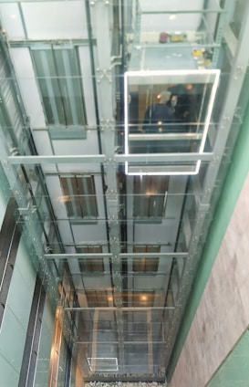 A glass elevator shaft extending vertically with a view upwards. A glass-walled elevator cabin is visible with two people inside. The structure is predominantly metallic and transparent, surrounded by glass walls, allowing visibility of the internal workings and multiple elevator doors on each floor.