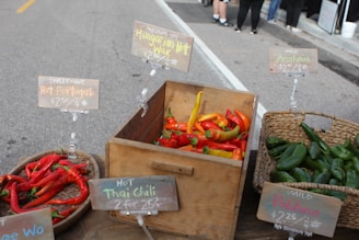A selection of fresh peppers displayed in baskets and a wooden crate at a market stall. The varieties include Hot Portugal, Hungarian Hot Wax, Anaheim, Thai Chili, and Poblano, each with a sign indicating its name and price per pound. The peppers are vibrant in color, with hues of red, yellow, and green, and are arranged neatly in their respective containers.