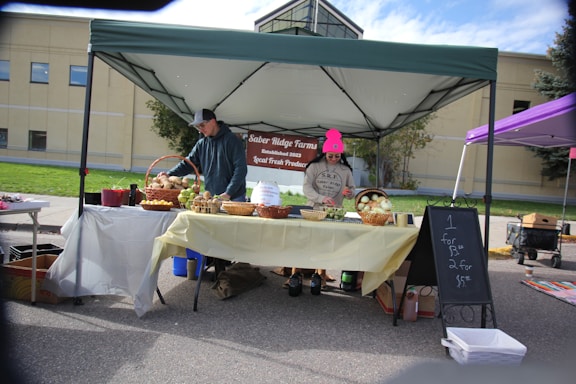 A small outdoor market stall with a green canopy displays fresh produce. The table under the tent is covered with a yellow cloth and features baskets filled with potatoes and onions. Two people are standing behind the table; one is looking at the produce while the other appears to be ready to assist customers. A chalkboard sign advertises prices for the produce. In the background, there is a building and another purple canopy visible.