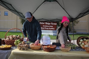 Two individuals are arranging an assortment of fresh produce on a table under a canopy, with a sign reading 'Saber Ridge Farms Established 2023 Local Fresh Produce' in the background. The table is covered with baskets containing various fruits and vegetables, such as potatoes, apples, lemons, and onions.