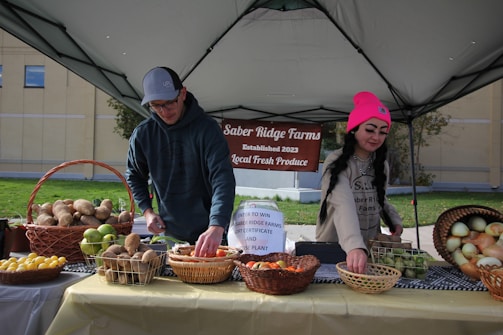 Two individuals are arranging an assortment of fresh produce on a table under a canopy, with a sign reading 'Saber Ridge Farms Established 2023 Local Fresh Produce' in the background. The table is covered with baskets containing various fruits and vegetables, such as potatoes, apples, lemons, and onions.