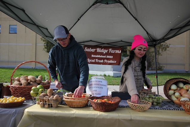Two individuals are arranging an assortment of fresh produce on a table under a canopy, with a sign reading 'Saber Ridge Farms Established 2023 Local Fresh Produce' in the background. The table is covered with baskets containing various fruits and vegetables, such as potatoes, apples, lemons, and onions.