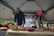 Two individuals are arranging an assortment of fresh produce on a table under a canopy, with a sign reading 'Saber Ridge Farms Established 2023 Local Fresh Produce' in the background. The table is covered with baskets containing various fruits and vegetables, such as potatoes, apples, lemons, and onions.