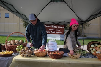 Two individuals are arranging an assortment of fresh produce on a table under a canopy, with a sign reading 'Saber Ridge Farms Established 2023 Local Fresh Produce' in the background. The table is covered with baskets containing various fruits and vegetables, such as potatoes, apples, lemons, and onions.