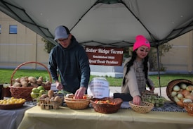 Two individuals are arranging an assortment of fresh produce on a table under a canopy, with a sign reading 'Saber Ridge Farms Established 2023 Local Fresh Produce' in the background. The table is covered with baskets containing various fruits and vegetables, such as potatoes, apples, lemons, and onions.
