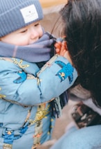 A young child is warmly dressed in a blue winter coat with colorful animal patterns and a matching blue knit hat. They are also wearing a scarf, secured by an adult, suggesting a caring interaction.