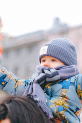 A young child wearing a blue knitted hat and a patterned winter jacket is dressed for cold weather. The child is also wearing a thick blue scarf wrapped around their neck. They appear to be sitting on someone's shoulders, as part of the adult's hair is visible at the bottom of the image. The background is softly blurred, suggesting an outdoor setting.