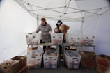 Two people are organizing packaged goods in large transparent bins under a white canopy tent. The bins are placed on tables, and there are cardboard boxes on the ground. The individuals are dressed warmly, suggesting a cold environment.