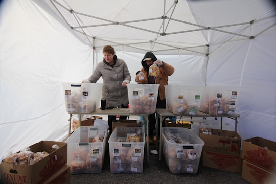 Two people are organizing packaged goods in large transparent bins under a white canopy tent. The bins are placed on tables, and there are cardboard boxes on the ground. The individuals are dressed warmly, suggesting a cold environment.