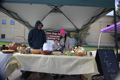 A market stall setup under a canopy, featuring a man, woman, and child standing behind a table filled with baskets of produce including potatoes, lemons, and onions. The woman is wearing a pink hat, and the man is wearing a dark hoodie. There is a sign advertising prices for the produce.