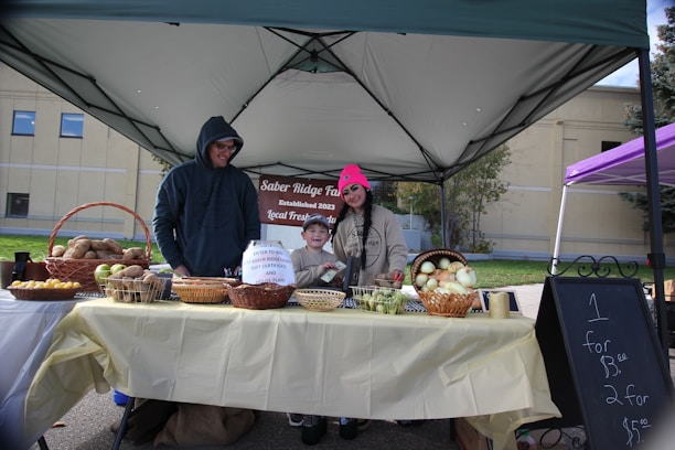A market stall setup under a canopy, featuring a man, woman, and child standing behind a table filled with baskets of produce including potatoes, lemons, and onions. The woman is wearing a pink hat, and the man is wearing a dark hoodie. There is a sign advertising prices for the produce.