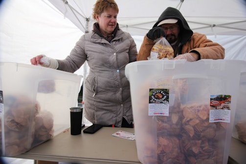 Two people are standing under a white tent, filling plastic bags with what appears to be potato chips from large plastic containers. Both individuals are wearing winter coats, suggesting it is cold. The table holds a few items including a cup, a phone, and a card. The containers have labels with branding.