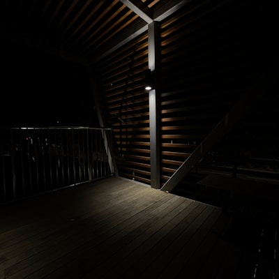 A wooden deck illuminated by a single overhead light, casting shadows on the slatted walls and floor. The area is enclosed by a railing on one side, and the light creates a dramatic play between light and shadow.