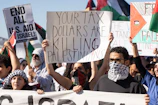 A group of people are gathered in a protest, holding various signs with messages related to political demands. Some signs read 'Your tax dollars are killing Palestinians' and 'End all U.S. aid to Israel'. Many individuals have their faces covered with scarves, and flags with distinct patterns can be seen in the background.