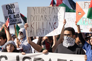 A group of people are gathered in a protest, holding various signs with messages related to political demands. Some signs read 'Your tax dollars are killing Palestinians' and 'End all U.S. aid to Israel'. Many individuals have their faces covered with scarves, and flags with distinct patterns can be seen in the background.